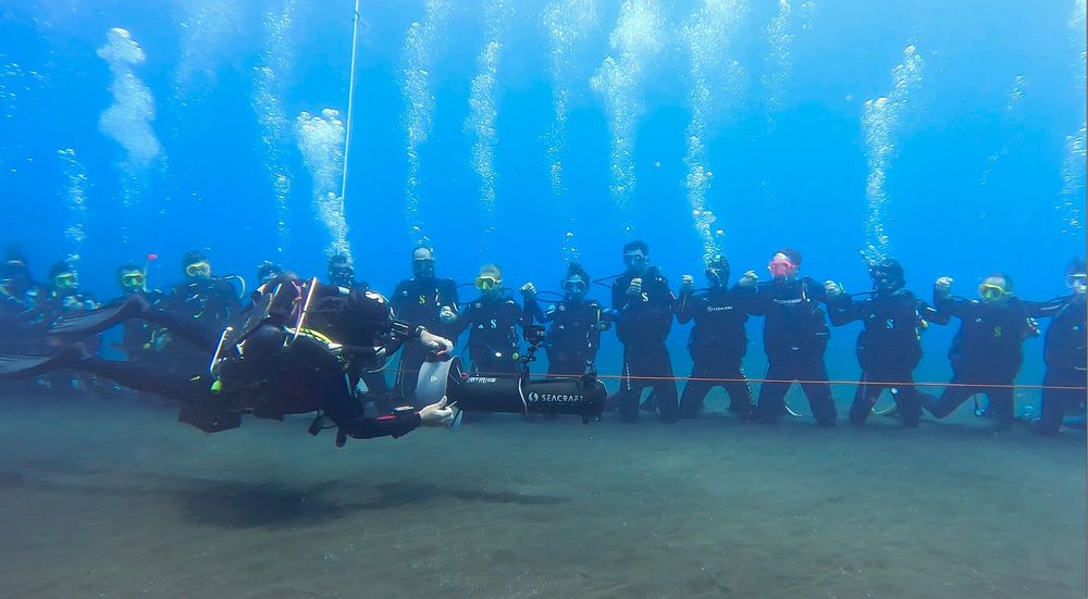 Des plongeurs côte à côte, passés en revue par un plongeur en scooter sous-marin.